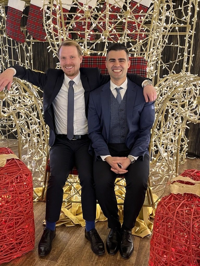Two men in dark suits sit side by side on a bench decorated with Christmas fairy lights, smiling at the camera.